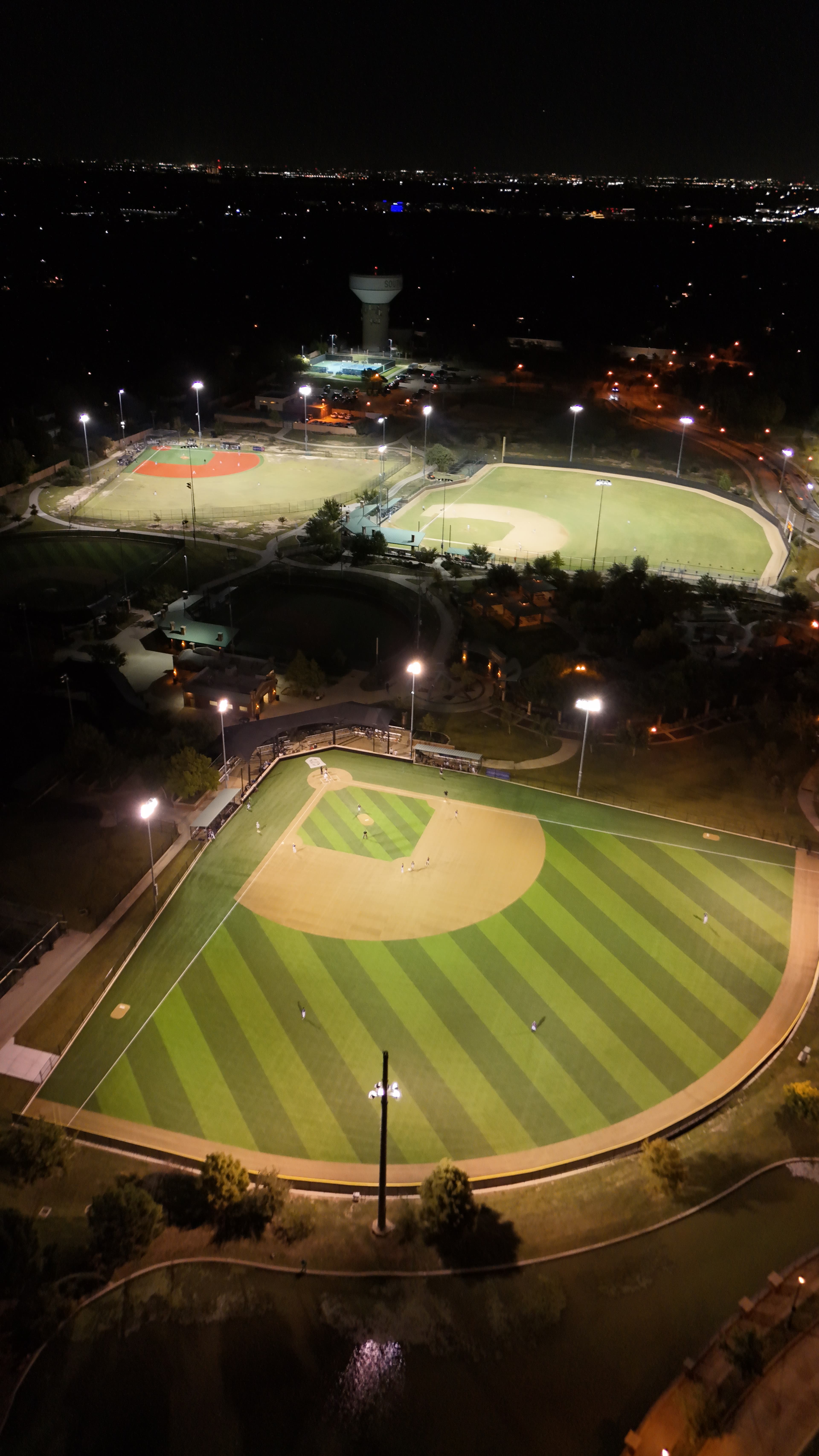 Night aerial drone photo of three baseball diamonds lit by stadium lights with city lights visible in the background