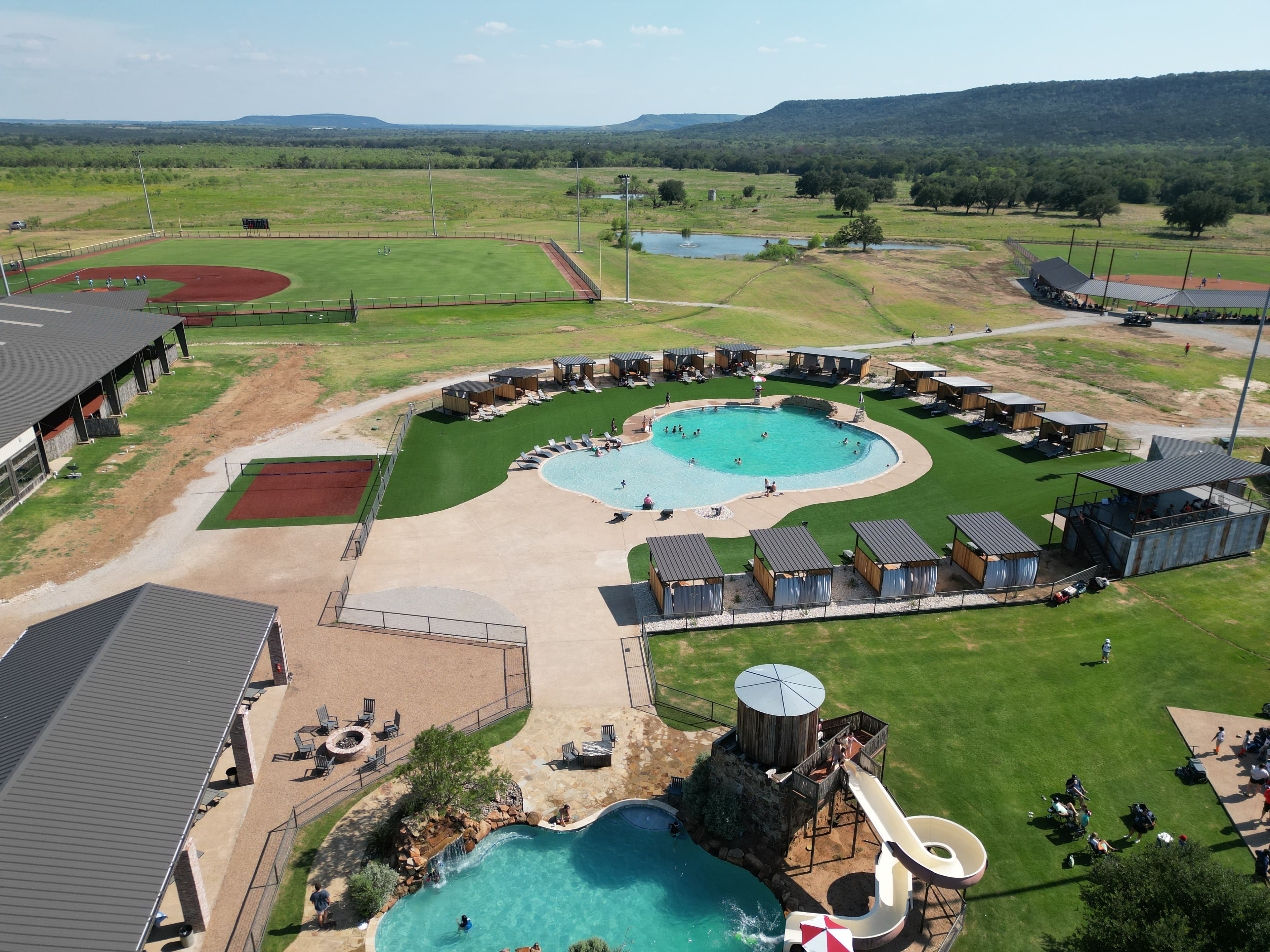 Aerial view of Texas Hill Country sports resort with pool, cabanas, baseball fields, and Palo Pinto mountains in background