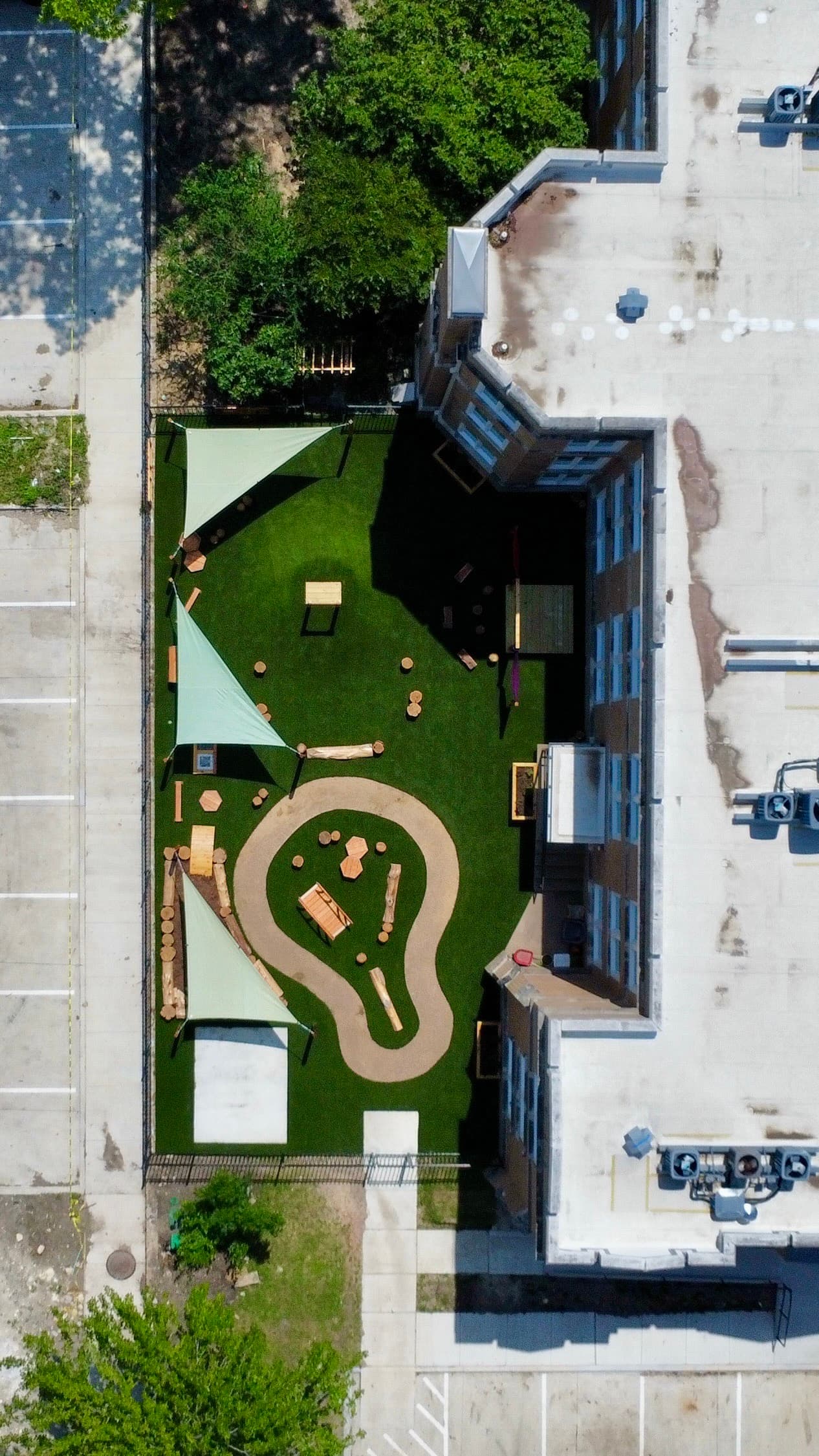 Overhead aerial of school courtyard playground with green turf, nature play path, shade sails, and urban surroundings