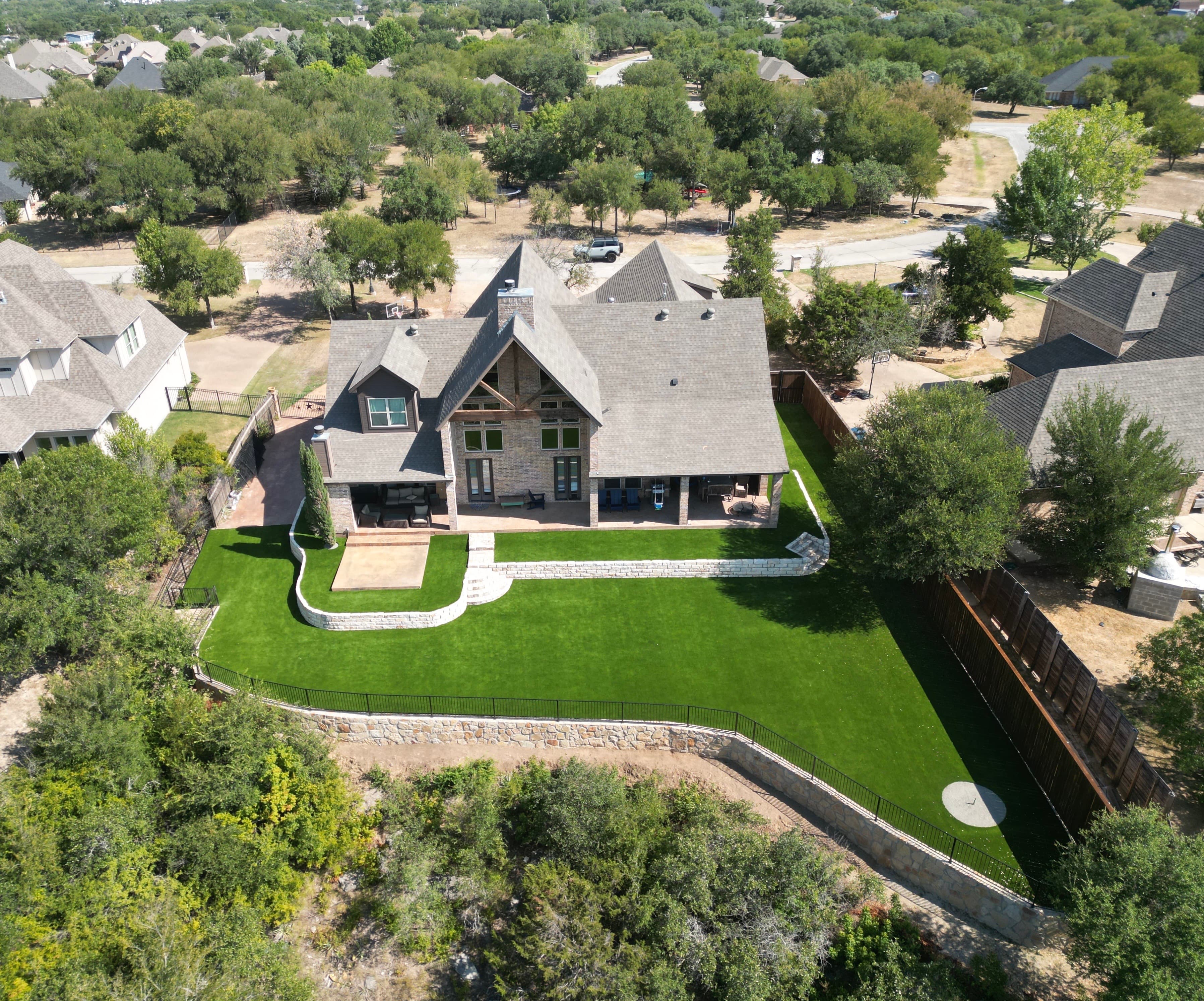 Aerial drone photo of custom home with lush green lawn, stone retaining walls, and putting green in Fort Worth suburb